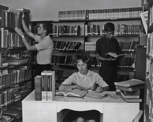 Vintage black and white photo, 3 students reading or interacting with books in a library