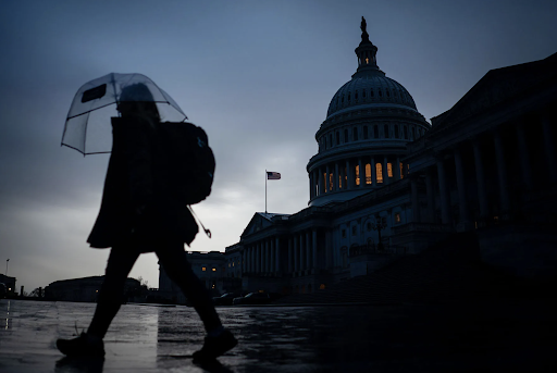 Photo of person walking in the rain with an umbrella, the Capitol Building darkened in the background
