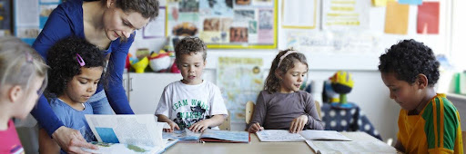 Photo of a teacher leaning over a student, assisting them, with other students reading around the table in a classroom