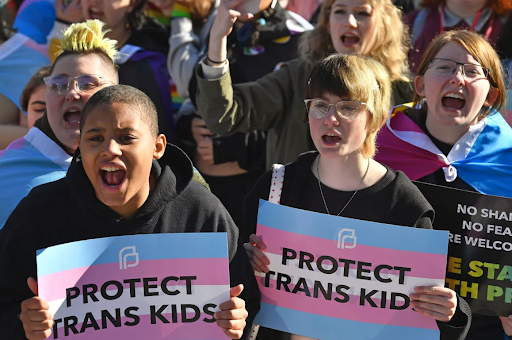 Photo of protesters holding signs of the trans pride flag with the message 