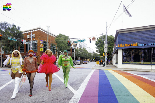 Photo of drag queens walking across the street of city scene, a rainbow flag painted on the ground to their right