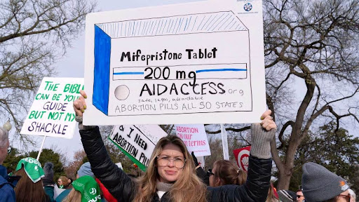Photo of a protester holding a poster of a hand-drawn box of Mifepristone
