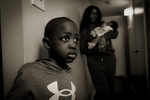 Black and white (sepia) photo of a young boy at the foreground, a mother holding a baby, seen blurry in the background