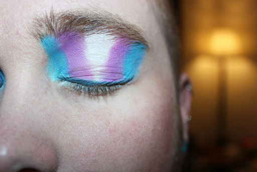 Photo: close-up of a person's face, one eye closed and full eyelid painted in the colors of the trans rights flag