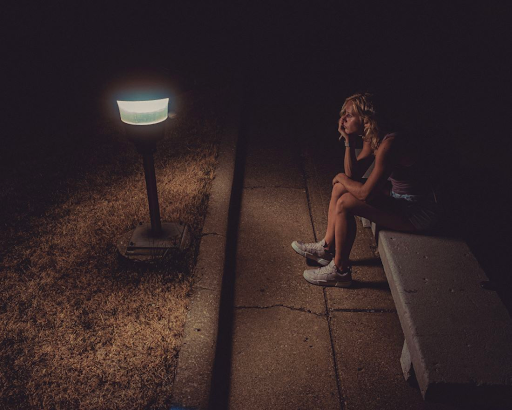 A young person sits on a park bench below a dim lamppost, posing in heavy thought.