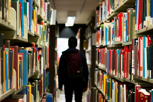 Photo: a person stands with tall shelves of library books on either side of them.
