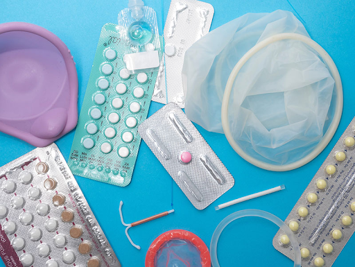 A photo of various contraceptive methods on top of a blue background. Methods featured include a diaphragm, birth control pills, an IUD, internal and external condoms and a Nuvaring. 
