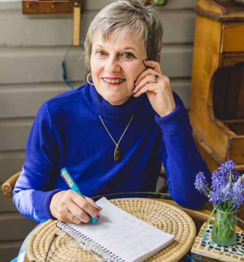 Photo of The Reverend Dr. Beverly Dale, wearing a royal blue turtleneck, sitting at a table, writing on a notepad placed on a round straw mat, a vase of vibrant violet flowers to her right.
