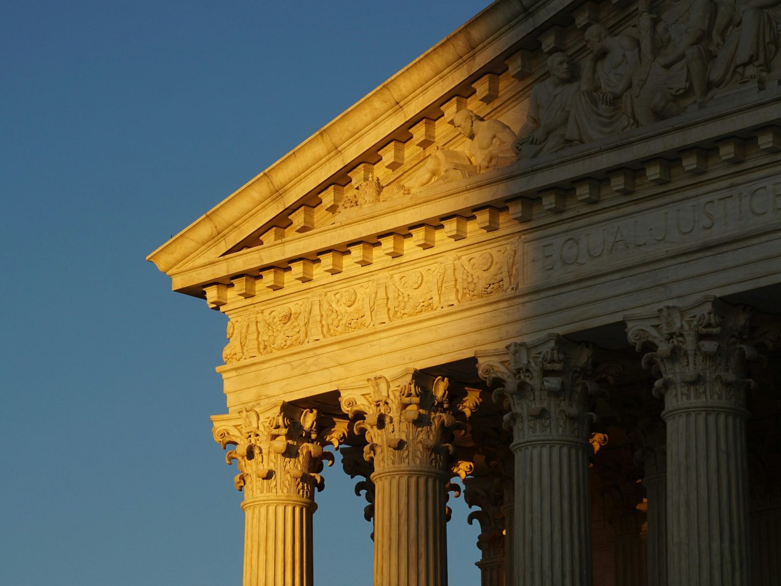 A photo of the upper corner of the Supreme Court Building. There is a shadow over the right side of the image. A gray-blue sky is the background. 